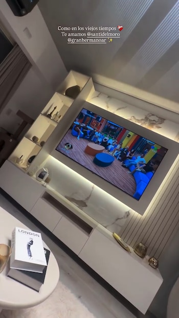 Una televisión de pantalla plana empotrada en una pared moderna, mostrando a los exconcursantes de Gran Hermano sentados en un sofá de colores dentro de la casa