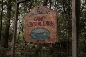 Un cartel de madera con la inscripción 'Welcome to Camp Crystal Lake' y 'Est. 1935' cuelga de un poste de madera en un denso bosque de árboles verdes