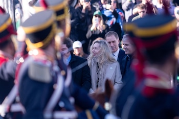 Primer plano de uniformes ceremoniales desenfocados. En foco, una mujer de cabello rubio entre dos hombres en ropa civil. Detrás, una multitud con teléfonos alzados