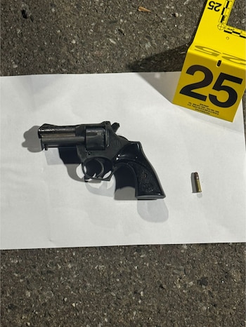 Un revólver negro, una bala dorada y un marcador de evidencia amarillo con el número 25 sobre una hoja de papel blanco en un suelo de asfalto