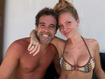 Un hombre y una mujer sonrientes posan juntos frente a una pared blanca, ella viste un traje de baño tejido
