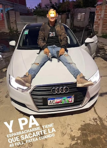 Hombre joven con la cara censurada, sentado sobre el capó de un Audi blanco, vistiendo jeans rotos y chaqueta, sosteniendo dos pistolas, en un entorno urbano