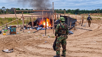 Un hombre con uniforme de camuflaje de espaldas frente a un campamento en llamas con postes de madera y lonas negras en un área arenosa.