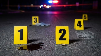 Una escena de crimen de noche muestra cinco marcadores de evidencia amarillos numerados en el asfalto, con sangre, un casquillo y una huella.