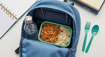 Mochila universitaria azul abierta con recipiente de comida de arroz con carne, botella de agua, tenedor y cuchara verdes, agenda y libros en el fondo.