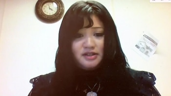 Mujer joven de cabello oscuro largo y flequillo, vistiendo una blusa negra, mira hacia adelante. Al fondo hay una pared blanca, un reloj y un papel
