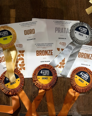 Primer plano de tres certificados de "4° Concurso de Queijos e Produtos Lácteos" con rosetas de oro, plata y bronce sobre una mesa de madera oscura