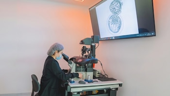 Una profesional de laboratorio con mascarilla opera un microscopio junto a un monitor que exhibe una imagen celular magnificada