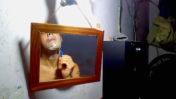 Reflejo en un espejo de un hombre afeitándose el cuello con una navaja azul, con espuma blanca y manchas de sangre. El espejo tiene un marco de madera y está colgado