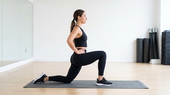 Una mujer con ropa deportiva negra realiza un estiramiento de flexores de cadera en posición de media rodilla sobre una colchoneta gris en un gimnasio.