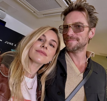 Una mujer rubia con collar y un hombre barbudo con gafas de sol posan en un primer plano de selfie, con un fondo oscuro y blanco