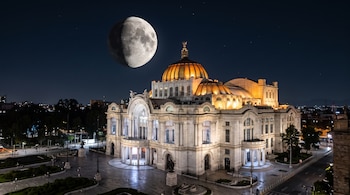 Palacio de Bellas Artes iluminado con luces cálidas bajo un cielo nocturno con la Luna y una explanada vacía al frente.