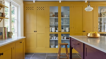 Interior de una cocina con gabinetes amarillos, isla morada con encimera de mármol, ventana, taburete de madera, vajilla y vasos, y suelo de baldosas oscuras.
