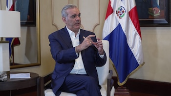 Luis Abinader, con traje azul y camisa blanca, sentado y gesticulando con las manos frente a la bandera dominicana y un cuadro