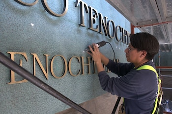 Un hombre con gafas y arnés de seguridad usa una herramienta eléctrica sobre letras doradas en una pared texturizada. Se lee parte de la palabra 'TENOCHTITLAN'
