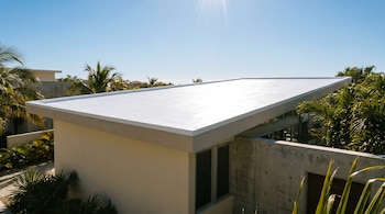 Vista exterior de una casa moderna con techo plano blanco reflectivo, paredes claras y una pared de concreto. Ventanas oscuras, palmeras, vegetación tropical y cielo azul.