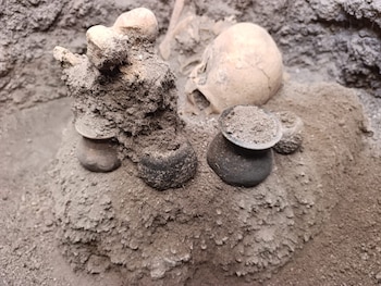 Restos óseos humanos, incluyendo un cráneo y huesos largos, junto a vasijas de cerámica cubiertas de tierra en un yacimiento arqueológico