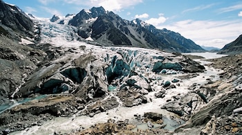 Glaciar con grietas de hielo azul y rocas expuestas junto a un río de agua de deshielo que fluye entre montañas bajo un cielo azul.