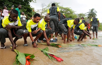 Grupo de hombres con uniformes deportivos, algunos agachados, colocando ramos de hojas y flores rojas en la orilla de una playa arenosa