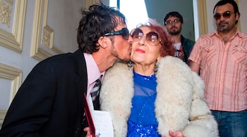 Un hombre con traje oscuro y gafas de sol besa en la mejilla a una mujer mayor con vestido azul, abrigo de piel y gafas de sol
