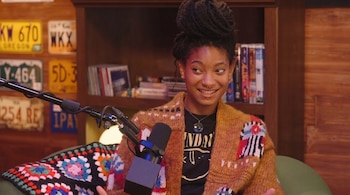 Willow Smith con peinado alto, suéter tejido y varios piercings sonríe frente a un micrófono de estudio