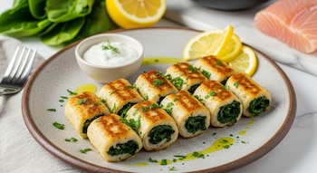 Diez rollos de merluza rellenos de espinaca, dorados y con perejil, se sirven en un plato de cerámica con salsa, limón y gotas de aceite.