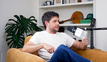 Niall Horan sentado en un sillón mostaza, con camiseta blanca y jeans, mirando a la derecha junto a un micrófono y una tarjeta 'And The Writer Is...'