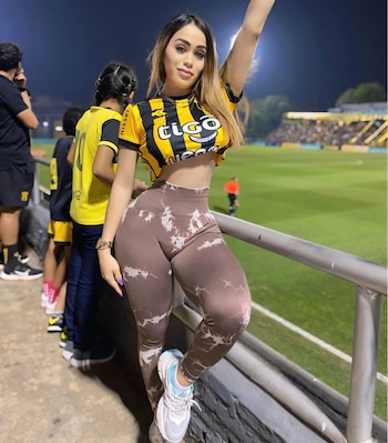 Marisol Olmedo, una mujer joven, posa sonriente en un estadio de noche, vistiendo una camiseta de rayas amarillas y negras y leggings estampados, con su brazo levantado