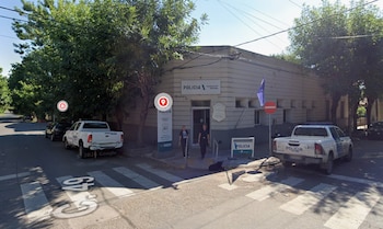 Edificio de una comisaría con cartel institucional, dos personas en la entrada y dos camionetas blancas aparcadas en la calle