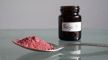 Un montón de polvo rosa rojizo brillante en una cuchara de laboratorio de acero, sobre una mesa de vidrio, con un frasco etiquetado 'Rojo 3' desenfocado detrás.