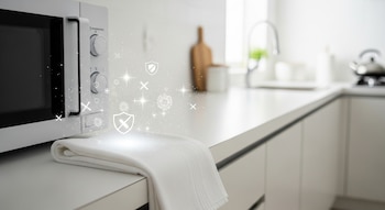 Un microondas blanco con controles, un trapo de cocina blanco doblado, una encimera blanca y símbolos gráficos de limpieza, virus y escudos.