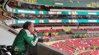 Hombre sentado de perfil con camiseta verde en un palco de estadio. En las pantallas se lee 'Copa Mundial de la FIFA 2026' y 'Mexico City'.