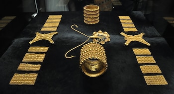 Conjunto de piezas de oro exhibidas sobre una superficie oscura que incluye ocho placas rectangulares, dos ornamentos, dos brazaletes y un collar