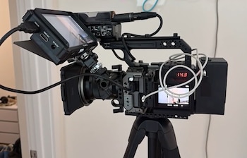 Cámara de video profesional negra sobre trípode. Cuenta con monitor externo, grabador de audio, cables y pantalla trasera con dígitos rojos