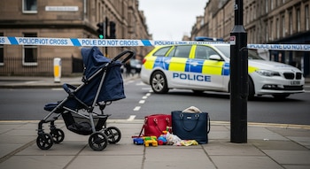Cochecito de bebé azul, dos bolsos (rojo y azul), juguetes de niños, una prenda de tela, cinta policial y patrulla en la calle con edificios de fondo.