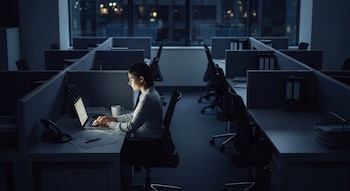 Una mujer sentada en un cubículo de oficina oscuro, trabajando en una laptop iluminada por la pantalla. El resto de los cubículos están vacíos.