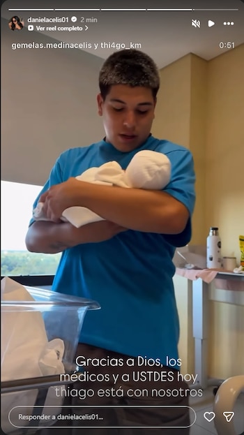 Hombre con camiseta azul sostiene en brazos a un bebé envuelto en una manta blanca dentro de una habitación