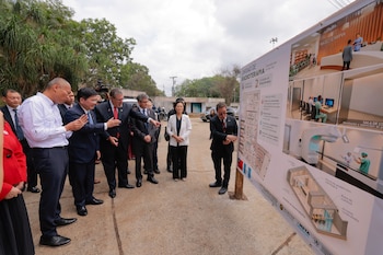 Un grupo de hombres en traje formal examina un cartel informativo con planos arquitectónicos e imágenes de instalaciones hospitalarias