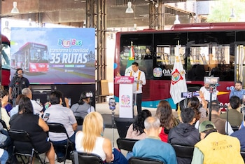 Hombre en podio blanco habla ante audiencia; detrás, pantalla con "BinniBus 35 RUTAS" y autobús rojo, y un autobús real estacionado