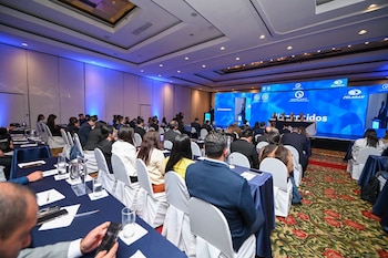 Personas sentadas en mesas con manteles azules dentro de una sala de conferencias frente a un escenario con pantallas LED
