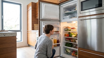 Vista posterior de una mujer agachada frente a un refrigerador abierto en una cocina moderna. El congelador tiene escarcha, mientras el refrigerador muestra alimentos organizados.