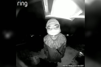 Grabación en blanco y negro de cámara Ring. Una persona con disfraz de payaso, gorro y gafas sostiene un objeto oscuro