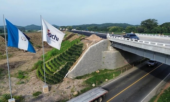 Vista aérea de una nueva autopista con un paso elevado de concreto en construcción. Banderas de Guatemala y de una empresa ondean en primer plano, con vehículos circulando