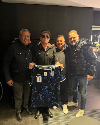 Cuatro hombres sonríen para la cámara en un interior oscuro; el hombre central sostiene una camiseta de fútbol de Argentina negra y azul con el número 10