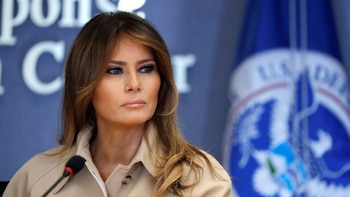 Retrato de Melania Trump, de tez clara y pelo castaño largo, luciendo un abrigo beige. Un micrófono en primer plano y un fondo azul con un logo borroso son visibles
