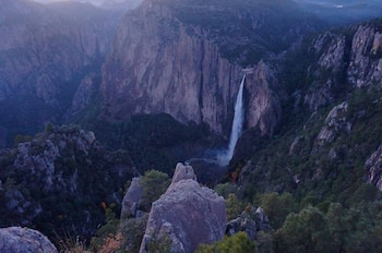 Paisaje montañoso con una cascada de agua cayendo por una pared rocosa, rodeado de picos escarpados y densa vegetación de árboles