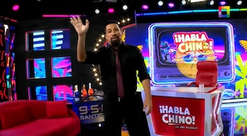 Aldo Miyashiro, vestido de camisa negra y corbata, saluda con la mano levantada en un estudio de televisión con luces de neón y el logo "¡Habla Chino!"