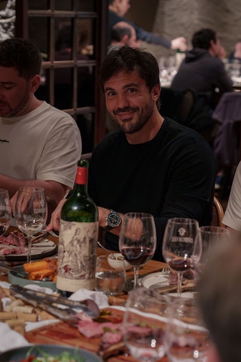 Un hombre sonríe sentado a una mesa con una botella de vino, copas con vino tinto, platos con carne asada y otros comensales borrosos
