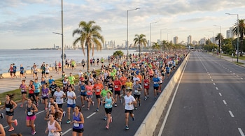 Vista aérea de cientos de personas corriendo en una carrera por un vial costero ancho, con el río, palmeras y el horizonte de la ciudad al fondo.