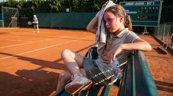 Una niña sentada en un banco verde en una cancha de tenis de arcilla, se seca el sudor con una toalla y sostiene una botella de agua. Al fondo, otra jugadora y un marcador.